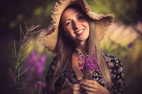 Etrafı mor çiçeklerle çevrili yaz şapkalı genç bir kadın.