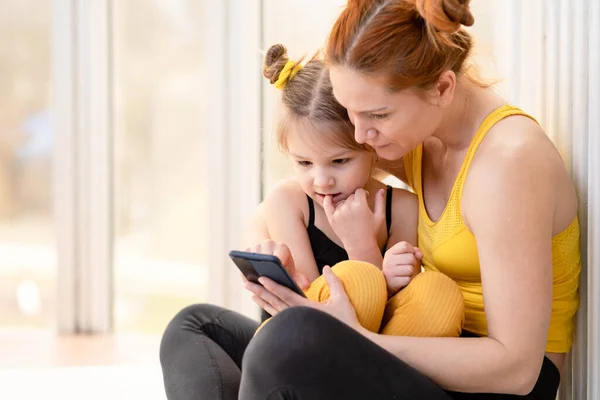 Young fit mom having loving time with her daughter, looking at a phone together