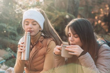 Two girls drinking hot tea from metal cups outdoor, camping