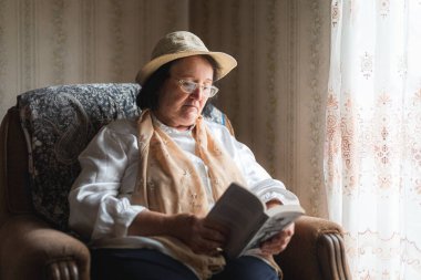 Yaşlı kadın kitap okuyor, pencerenin yanındaki sandalyede oturuyor.