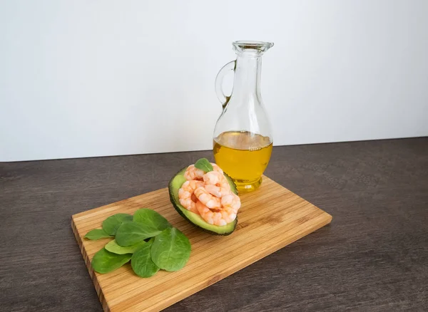 Minimalist conceptual composition with fresh green avocado, pink shrimps and transparent olive oil in a jug, salade leaves on a wooden cutting board, healthy food diet