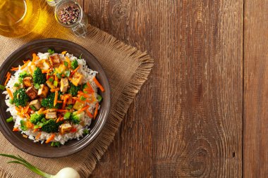 Pilav ve sebzeli soya peyniri. Kahverengi tabakta yemek çubuklarıyla servis ediliyor. Üst görünüm.