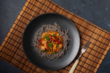 Siyah susamlı buğday eriştesi, tavuk ve sebzeli tavada kızartılmış. Üst Manzara. Siyah bir tabakta servis edildi..