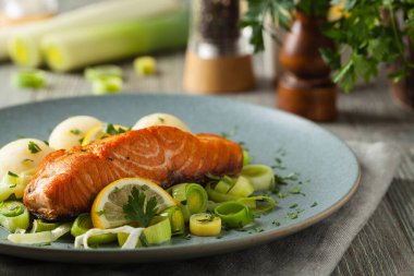 Bir porsiyon kızarmış somon, patates püresi ve pişmiş pırasa ile servis edilir. Ön manzara. Gri plaka, ahşap arka plan. 