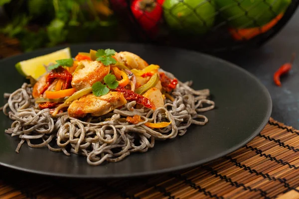 Wheat noodles with black sesame, fried in a wok with chicken and vegetables. Front view. Served on a black plate.