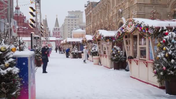 Plusieurs touristes marchent à travers la Foire du Nouvel An sur la Place Rouge un jour d'hiver lors d'une chute de neige. — Video