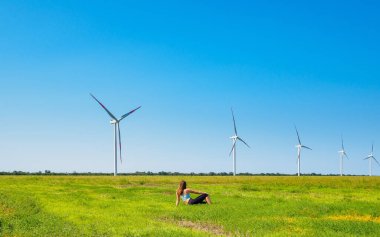 Rüzgâr türbinlerinin önündeki çayırda oturan genç bir kadının arka görüntüsü.. 
