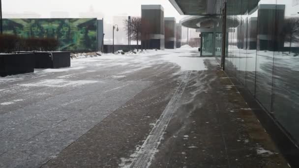 Nieve llevada por el viento se mueve a lo largo de la acera cerca de un edificio moderno desconocido con una fachada de vidrio. — Vídeo de stock