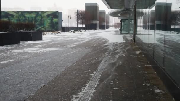 Vom Wind getragener Schnee bewegt sich auf dem Bürgersteig in der Nähe eines unbekannten modernen Gebäudes mit Glasfassade. — Stockvideo