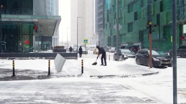 En servicearbetare skyfflar snö med en spade på en stadsgata under ett snöfall på en vinterdag. — Stockvideo