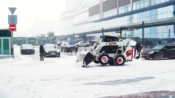Un pequeño cargador con un cubo elimina la nieve de una carretera en una calle de la ciudad — Vídeos de Stock