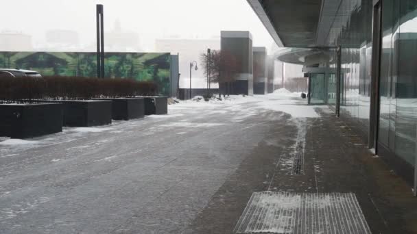 ガラスのファサードを持つ未知の近代的な建物の近くの歩道に沿って風が運ぶ雪. — ストック動画