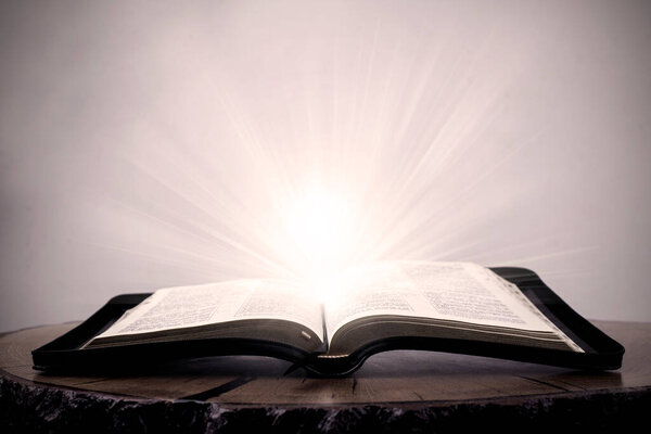 Открытая Библия. Большая книга Библии лежит на деревянном столе. В темноте. Свет сияет на книгу сверху. Свет выходит из книги