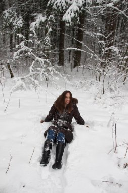 Kahverengi kürklü genç kadın kar altında oturuyor ve kış ormanında gülümsüyor.