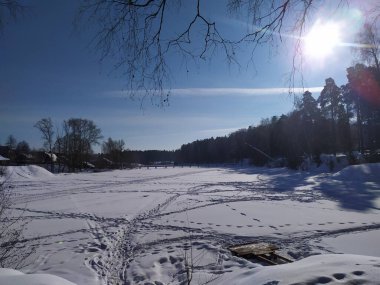 Mavi gökyüzünde kar, donmuş nehir, orman ve güneş olan kır manzarası