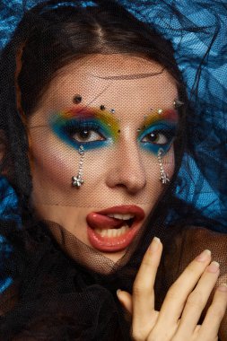 Moda yaratıcı makyaj. Güzel bir genç kadın portresi. Dili dışarı çıkmış. Taze bir ten ve parlak bir makyajla parlak mavi bir arka planda poz veriyor.