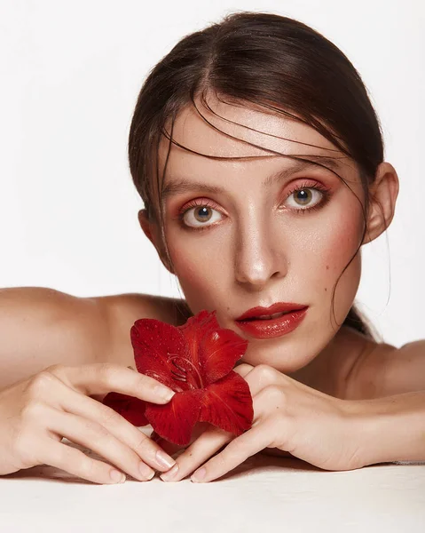 Yüzünün yanında kırmızı çiçekli güzel bir kadın çıplak omuzları açık pembe makyajlı bir cilt modeli. Beyaz arka planda cilt bakımı ve kaplıca konsepti. Kameraya bakan kadın.