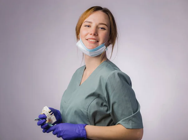 作業ツールと魅力的な女性歯科医の肖像画は 均一な医療面マスクとラテックス医療手袋を着用してください 灰色の背景にプラスチック製の入れ歯の歯とツールを保持する — ストック写真