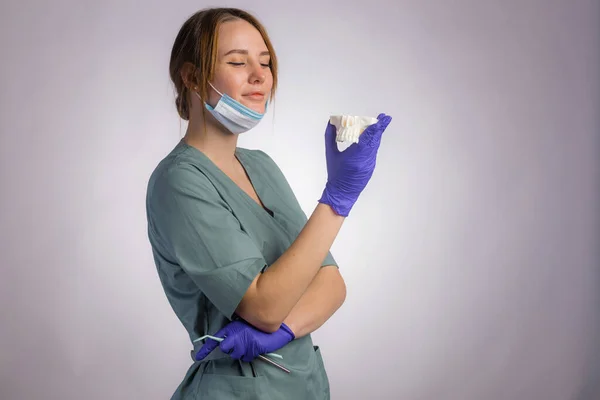 作業ツールと魅力的な女性歯科医の肖像画は 均一な医療面マスクとラテックス医療手袋を着用してください 灰色の背景にプラスチック製の入れ歯の歯を保持 — ストック写真
