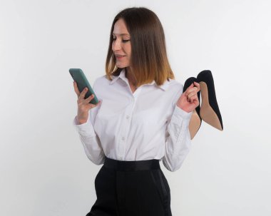 Mesai bitimi - dinlenme zamanı. İşten sonra yüksek topuklu ayakkabılarını çıkaran mutlu iş kadını, başarıyla tamamlanmış çalışmalarını kutluyor. Beyaz fotokopi alanında izole edilmiş telefonla haberleri paylaşıyor