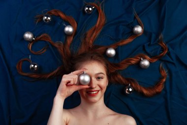 Saçının ucunda dekoratif noel toplarıyla mavi yatakta uzanmış ve bir topu gözlerinin yanında tutan tatlı kızıl saçlı genç bir kadın. Üst görünüm