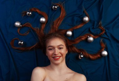 Saçının uçlarında dekoratif noel toplarıyla mavi yatakta yatan tatlı kızıl saçlı genç bir kadın. Üst görünüm