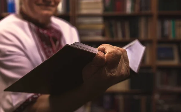 把手里拿着一本书的老手绑起来 穿着民间服装的老人在家里的图书馆里拿着一本书 — 图库照片