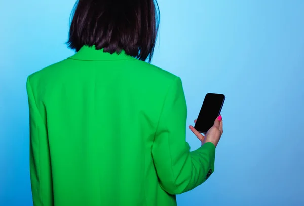 Close up woman holding phone with blank screen on blue background and copy space. Rear view woman in green blazer