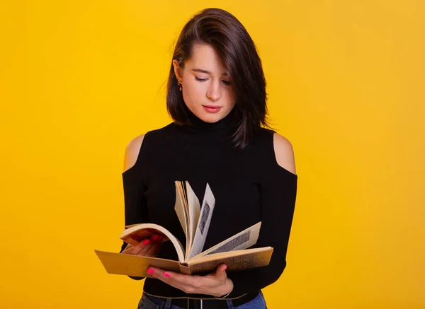 Pretty woman with a book in hands smiling at camera. Clever and excited lady holding open book. Studio shot isolated on yellow background