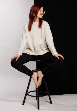 Siyah tayt ve beyaz kazakla poz veren büyük gözlüklü kızıl saçlı güzel bir manken. Yarı siyah beyaz arka planda bir sandalyede oturmak. Güzellik, moda konsepti. İş biçimi.