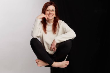 Siyah tayt ve beyaz kazakla poz veren büyük gözlüklü kızıl saçlı güzel bir manken. Yarı siyah beyaz arka planda bir sandalyede oturmak. Güzellik, moda konsepti. İş biçimi.