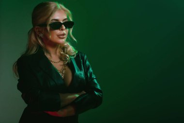 Studio fashion shot of the beautiful rock woman dressed in black leather jacket and black body lingerie, sunglasses on the face. Seductive lady in green light on a dark green background