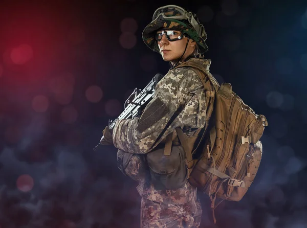 Woman army soldier in combat uniforms with assault rifle, plate carrier, goggles and backpack. Studio shot in smoke, dark background