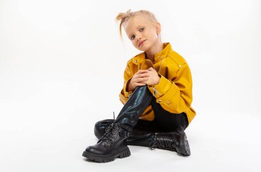 Pretty blonde little girl sitting in studio floor in a leather mustard jacket, black snake texture leggings and boots. High fashion, full length standing pose, isolated against a studio background