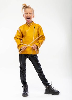 Rock and roll music concept. Girl musician, holding drum sticks in hands and shows her tongue. Dressed in a leather mustard jacket, black leggings and boots. Full length, istudio background
