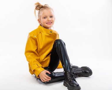 Pretty blonde little girl sitting in studio floor in a leather mustard jacket, black snake texture leggings and boots. High fashion, full length standing pose, isolated against a studio background