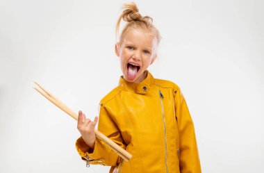 Rock and roll music concept. Girl musician, holding drum sticks in hands and shows her tongue. Dressed in a leather mustard jacket, black leggings and boots. Full length, istudio background