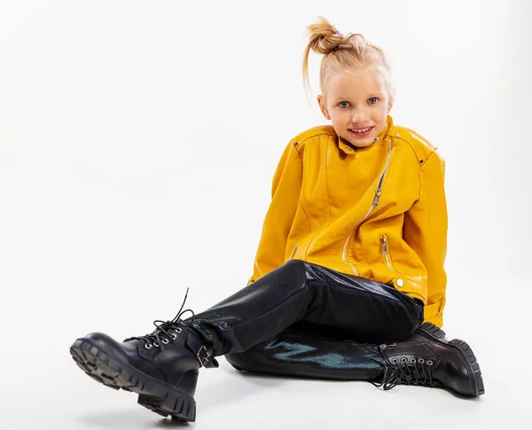 Pretty blonde little girl sitting in studio floor in a leather mustard jacket, black snake texture leggings and boots. High fashion, full length standing pose, isolated against a studio background