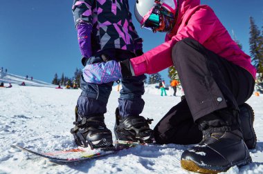 Anne kızına snowboard yapmayı öğretiyor. Güneşli kış günlerinde kayak merkezindeki kaykayı bağlamasına yardımcı olur. Seyahat tatili, manzara dağları....