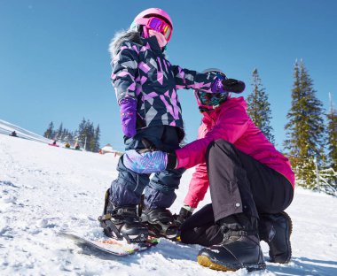 Anne kızına snowboard yapmayı öğretiyor. Güneşli kış günlerinde kayak merkezindeki kaykayı bağlamasına yardımcı olur. Seyahat tatili, manzara dağları....