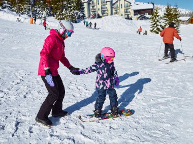 Anne kızına snowboard yapmayı öğretiyor. Güneşli kış günlerinde kayak merkezinde kayak yapan çocuklara yardım eden ve destek olan bir kadın. Seyahat tatili, manzara dağları geçmişi..