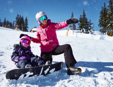 Annesi kızına snowboard yapmayı öğretiyor. Kadın oturur ve güneşli kış gününde kayak yamacında yol gösterir. Seyahat tatili, manzara dağları....