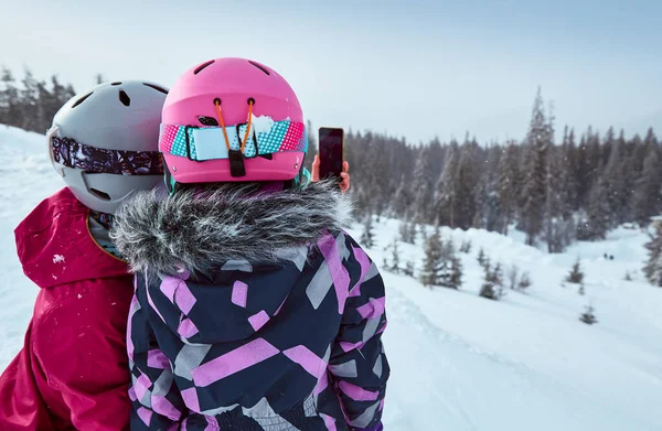 Back view of mother and daughter in ski equipment together taking selfies or video call in the mountains on ski resort at sunny winter day, travel vacation, or network coverage and connection concept.