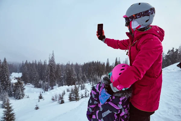 Back view of mother and daughter in ski equipment together taking selfies or video call in the mountains on ski resort at sunny winter day, travel vacation, or network coverage and connection concept.