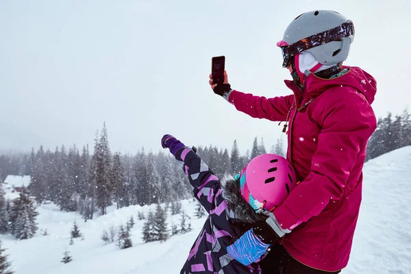 Back view of mother and daughter in ski equipment together taking selfies or video call in the mountains on ski resort at sunny winter day, travel vacation, or network coverage and connection concept.