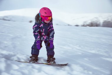 Güneşli kış gününde kayak merkezinde küçük bir snowboardcu kız. Spor kıyafetli, teçhizatlı çocuk portresi.