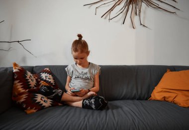 Oturma odasındaki kanepede oturan tatlı kız akıllı telefon kullanıp online oyunlar oynuyor. Telefonu tutup ekran cihazına bakıyorum. Çocuk kanepede oturuyor ve oturma odasında dinleniyor. Teknoloji konsepti.