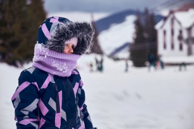 Kış tatilinin tadını çıkaran küçük gülümseyen kızın portresi. Kayak merkezinde kapüşonlu ve kürklü örülü şapka ve yüz kaplaması olan çocuk karlı arka planda. Karda oynayan kız