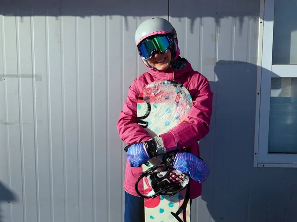 Winter sport woman posing with snowboard on the background of ski pass office. Woman snowboarder holding a snowboard outdoors and smiling at camera