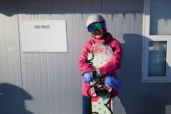 Winter sport woman posing with snowboard on the background of ski pass office. Woman snowboarder holding a snowboard outdoors and smiling at camera